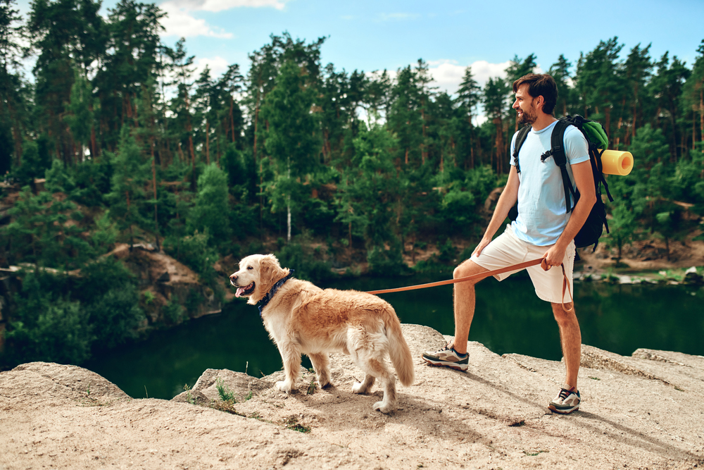 A man with a backpack and yoga mat stands on a rock ledge in a mountain for a hike