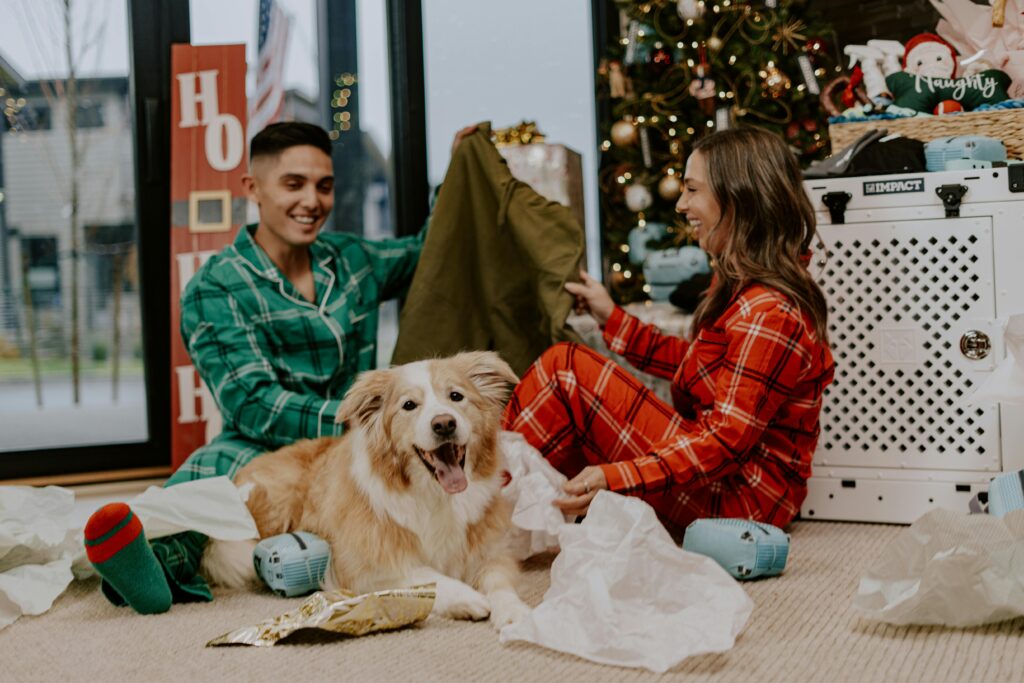 A couple in pajamas sits on the floor by a Christmas tree with a happy dog, unwrapping gifts and smiling.