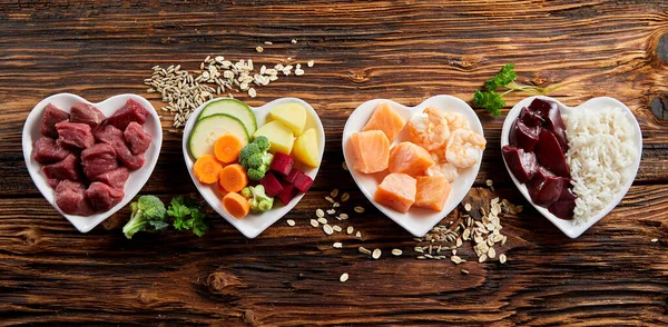 Four heart-shaped bowls with raw beef, mixed vegetables, salmon with shrimp, and rice with liver on a wooden surface, surrounded by scattered grains and herbs.