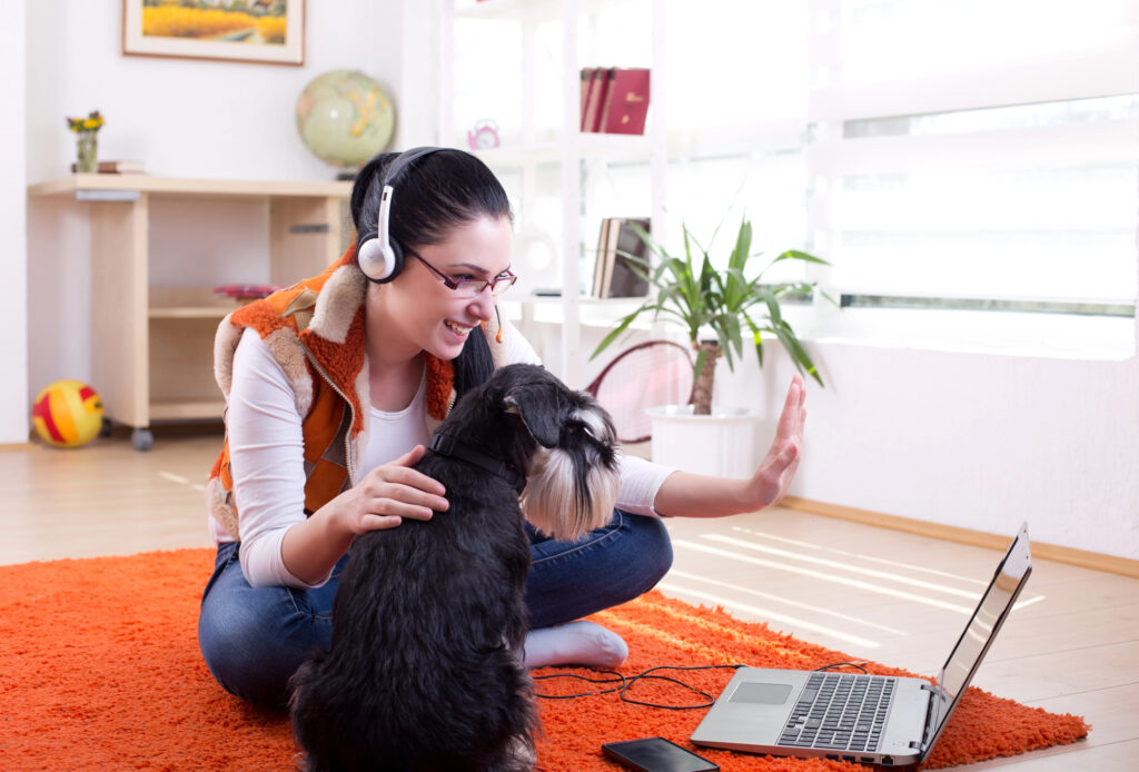 A woman wearing headphones sits on an orange rug with a dog, waving at a laptop during a video call in a bright room.