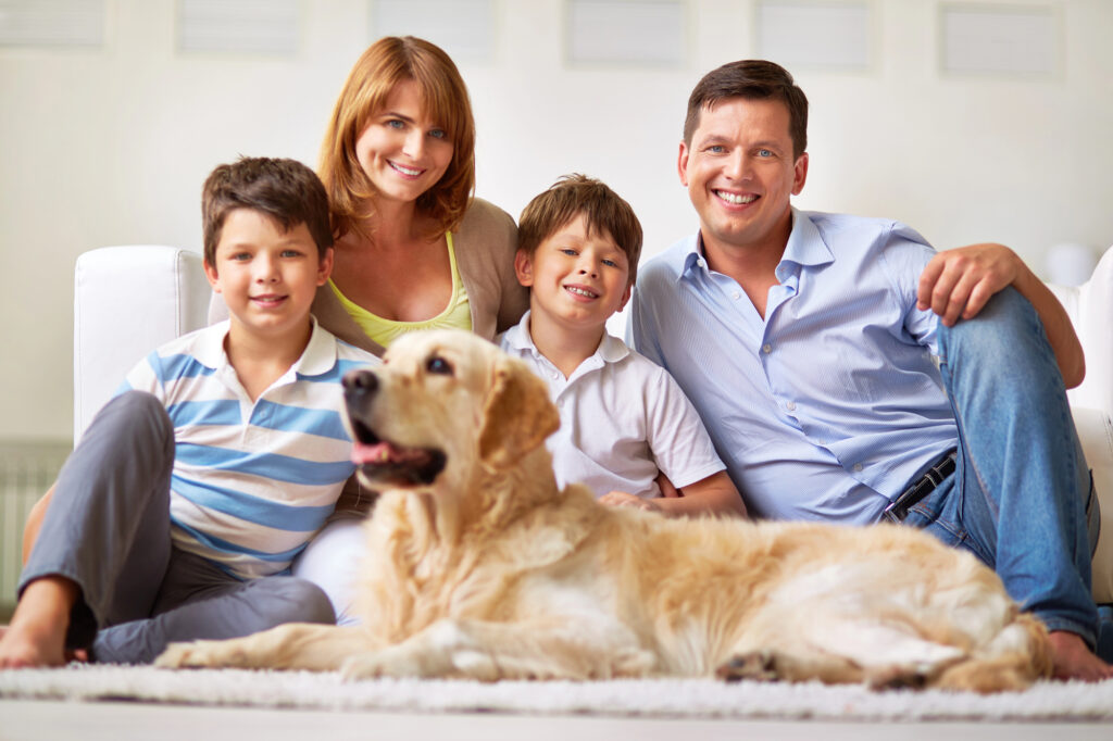 A family of four sits on a couch with a golden retriever lying in front of them, all smiling at the camera in a bright living room.