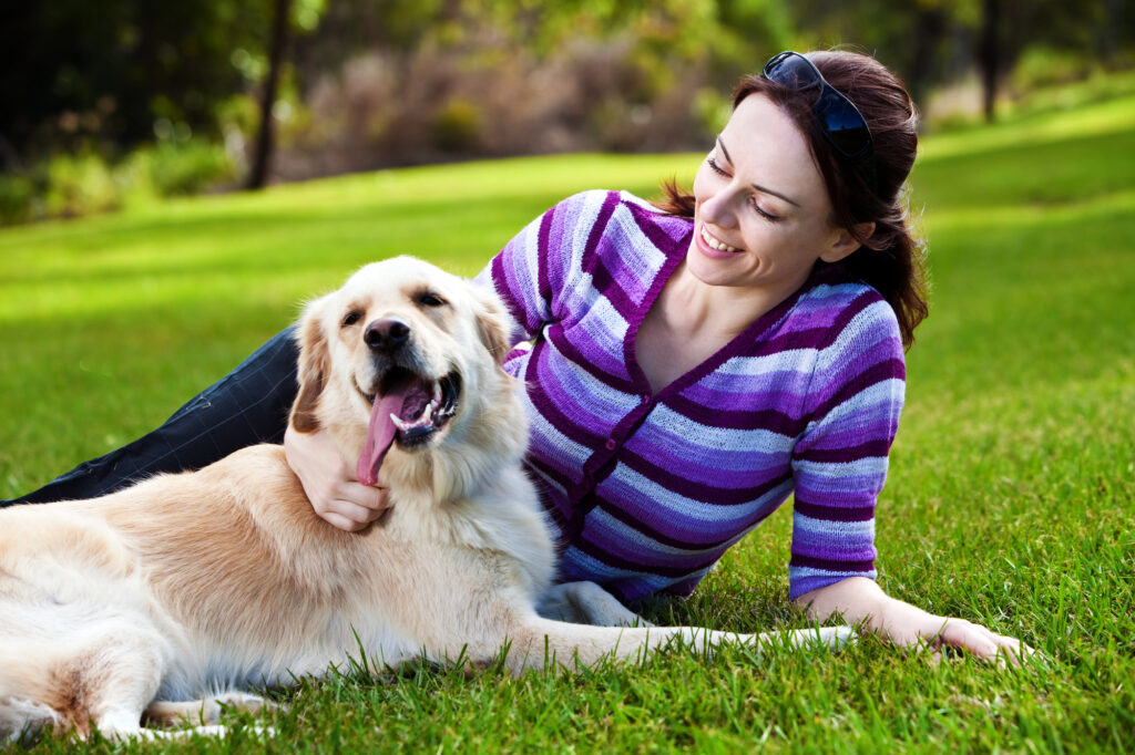 A woman lying on grass smiles while petting a golden retriever, who is lying beside her with its mouth open. The scene is outdoors in a park.