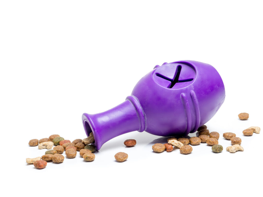 A purple rubber dog treat dispenser toy is tipped over with dry pet food spilling out onto a white surface.