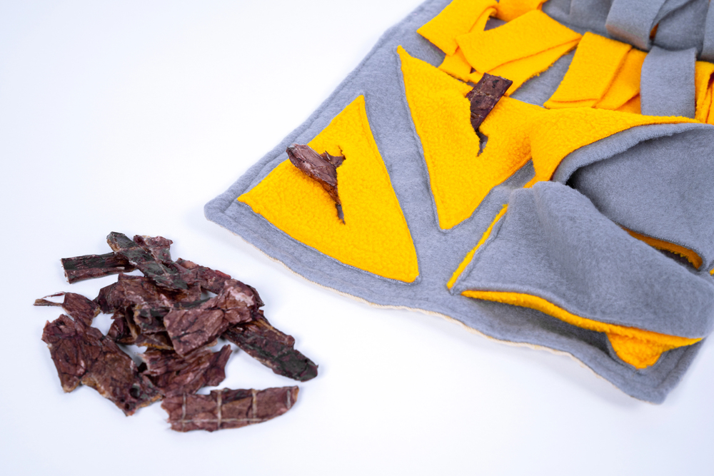 Gray and yellow fleece snuffle mat with several pieces of dried meat placed inside and more pieces scattered beside it on a white background.