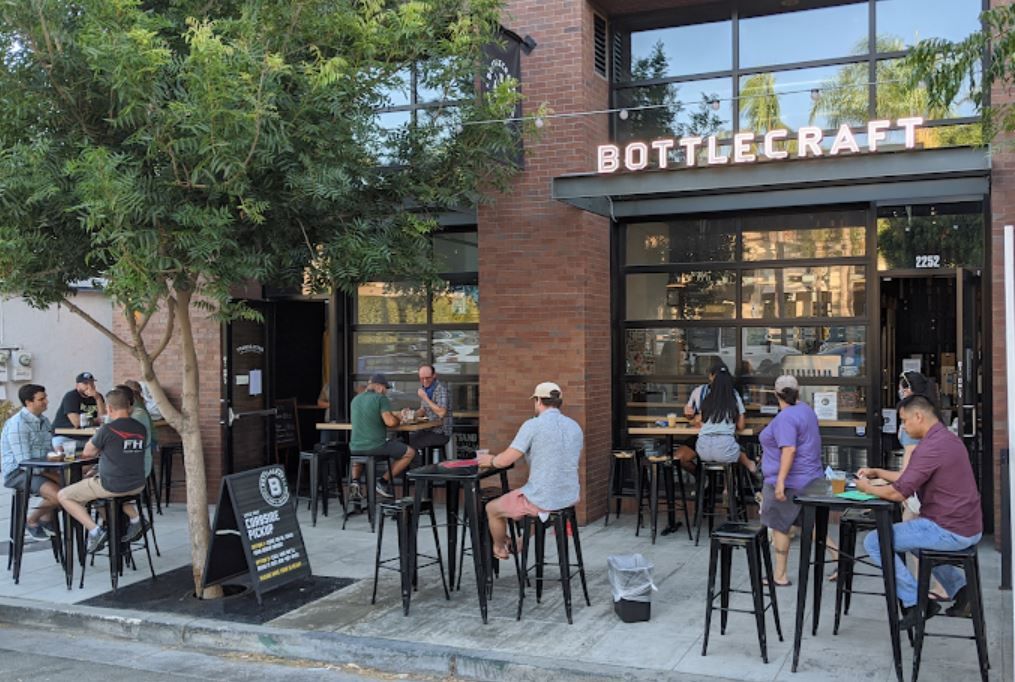People sit at outdoor tables in front of a bar called Bottlecraft on a sunny day, with trees providing some shade and a signboard displayed near the entrance.