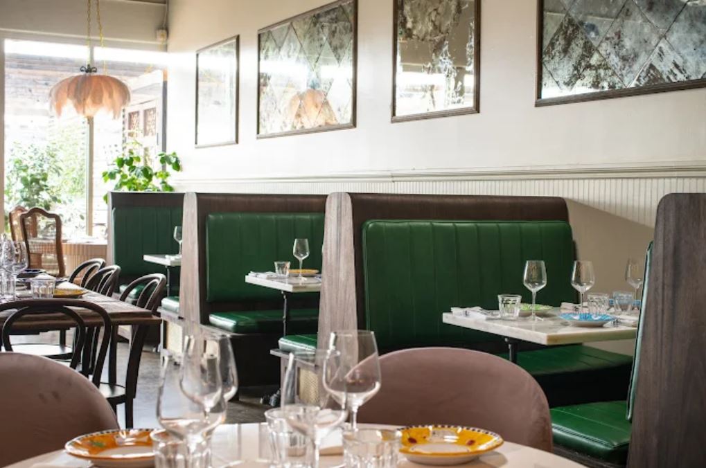 A restaurant interior with green booth seating, set tables with glasses and plates, framed mirrors on the wall, and natural light coming from the windows.