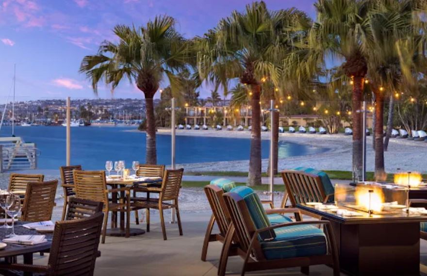 Outdoor restaurant patio with wooden tables and chairs overlooks a beach and marina, with palm trees, string lights, and a calm evening sky in the background.