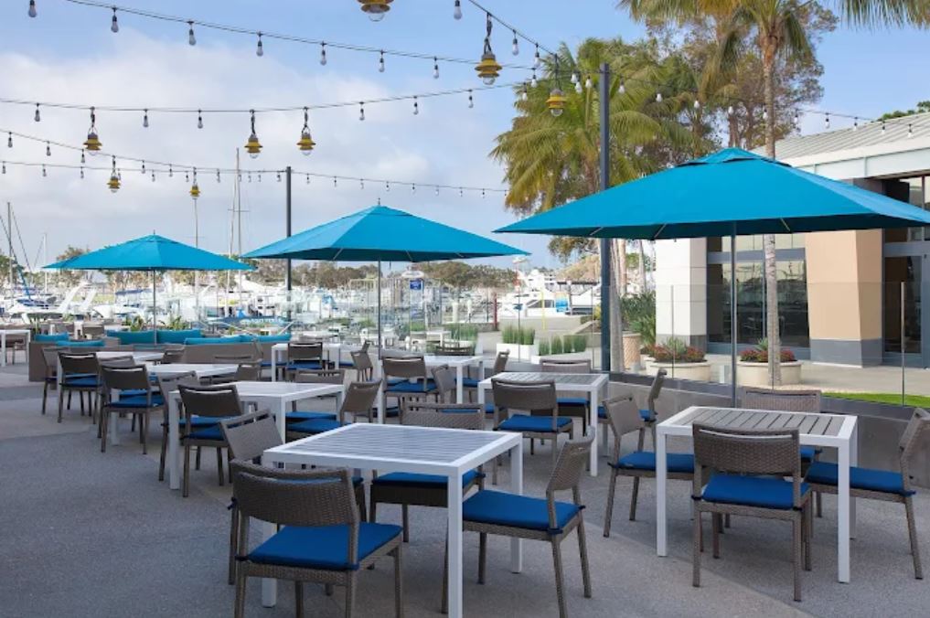 Outdoor patio with white tables, blue cushioned chairs, and blue umbrellas; string lights above and a marina with boats in the background.