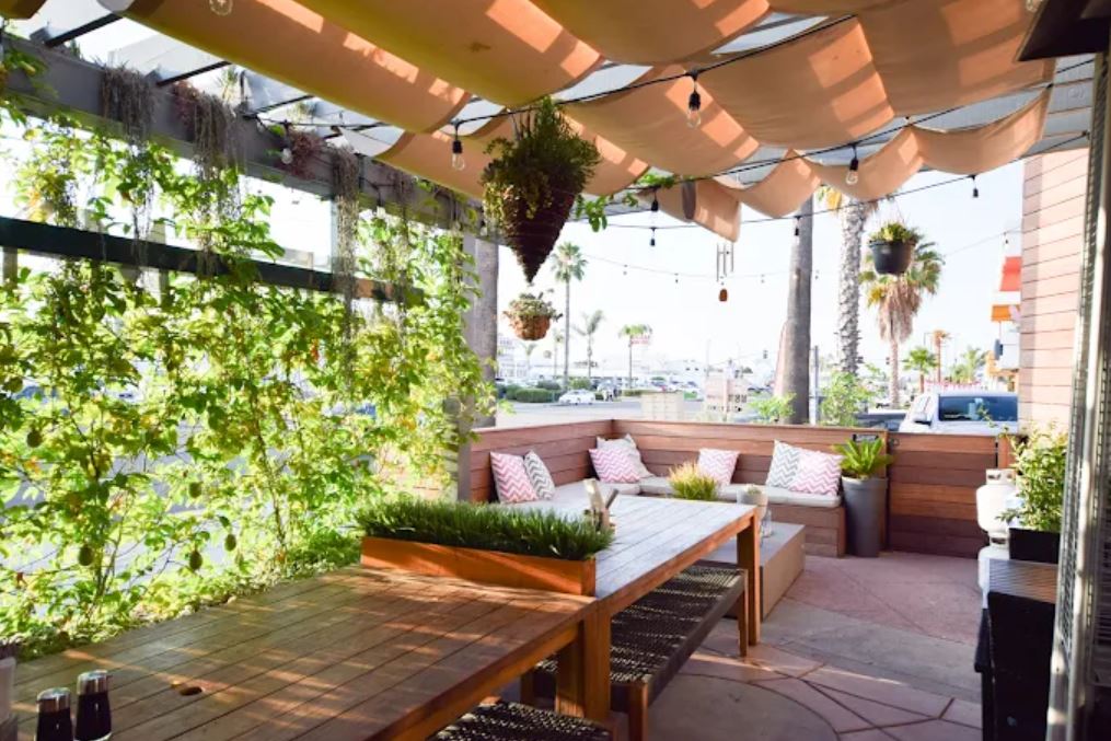 Outdoor patio with wooden tables, cushioned seating, hanging plants, string lights, and greenery on one side, under a beige canopy.
