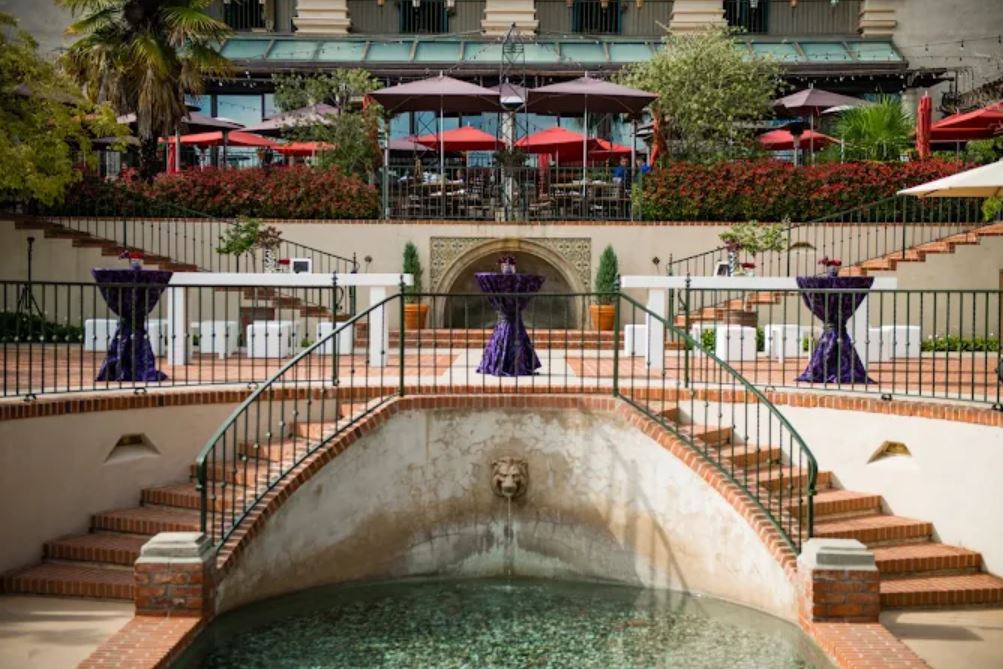 Outdoor terrace with red umbrellas, round tables covered in purple cloth, a small fountain, and ascending brick stairs surrounded by greenery and railings.