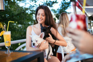 A woman sitting at an outdoor café smiles while holding two small dogs, one white and one black, with drinks and other people in the background.