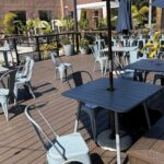 Outdoor dining area at Moniker Coffee in San Diego with empty blue metal tables and chairs on a wooden deck, surrounded by plants and shaded by umbrellas on a sunny day.