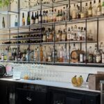 A bar counter with various liquor bottles, wine bottles, glasses, and hanging greenery above shelves labeled "Cocktails.