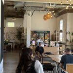 A person with a backpack walks into a spacious café with exposed ceilings, wooden tables, and a flower display in the background. Other people are seated and working.