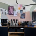 A coffee station with carafes, cups, condiments, and a poster on the wall advertising “Tues Trivia 6 PM.” Flyers hang above the counter on a wire grid.