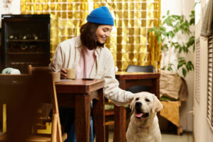 A woman in a blue beanie sits at a wooden table with a cup, smiling and petting a Labrador Retriever in a cozy coffee shop