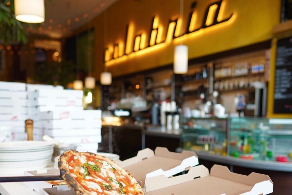 A pizza in an open box sits on a counter in a restaurant, with stacked pizza boxes and a lit "calabria" sign visible in the background.