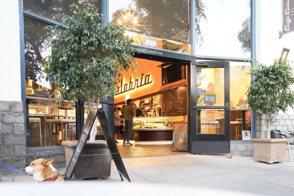 Two people stand inside a coffee shop with large windows, while a corgi on a leash sits outside near potted plants on the sidewalk.