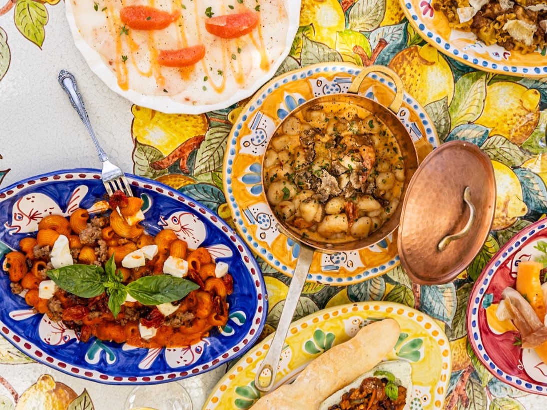A table set with colorful plates of Italian food, including pasta, gnocchi, breadsticks, and sliced tomatoes, on a decorative lemon-patterned tablecloth.
