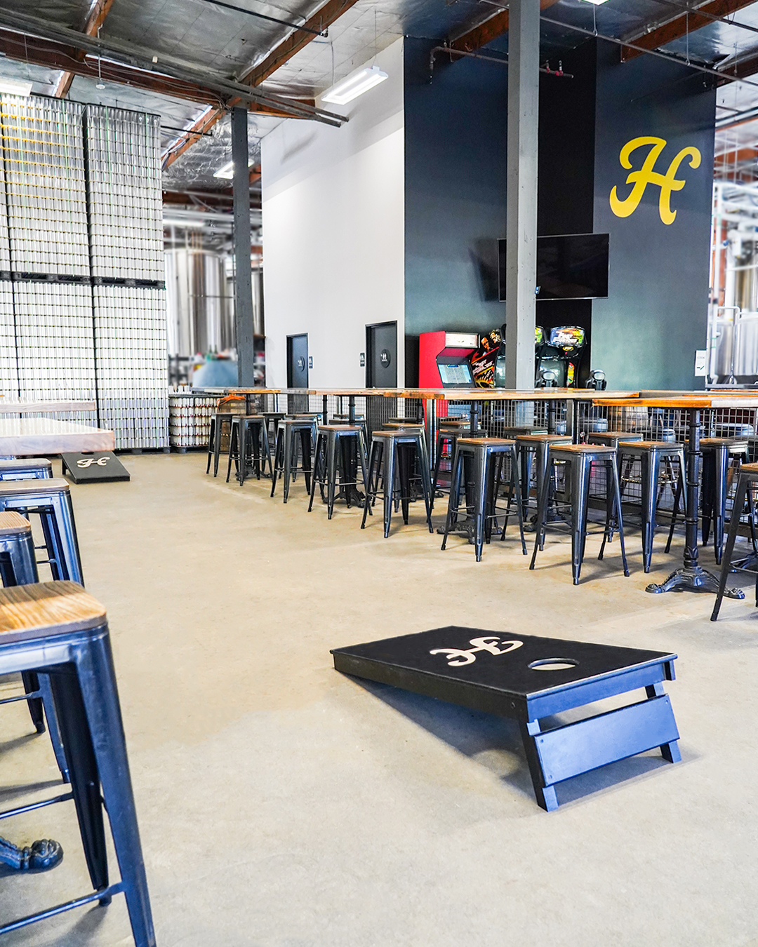 Spacious industrial bar with high tables and stools, arcade games, and a cornhole game set on the floor; large yellow "H" on a dark wall.
